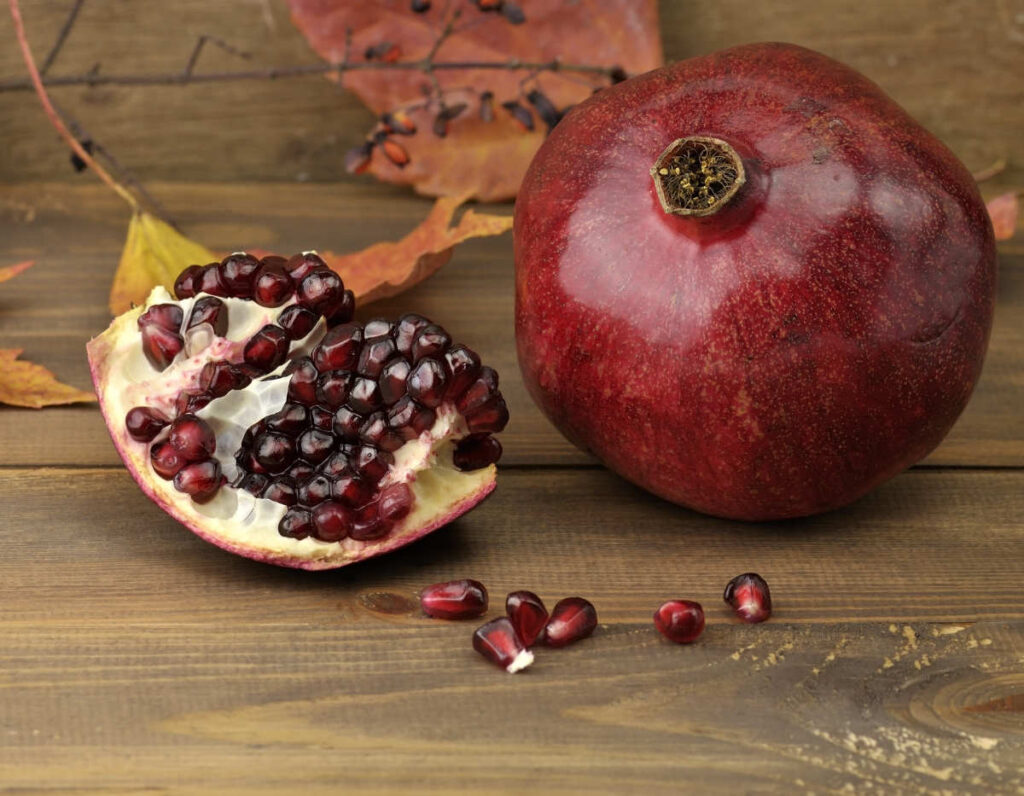 Ein ganzer und aufgeschnittener Granatapfel mit sichtbaren Kernen liegt auf einem Holztisch neben Herbstblättern.
