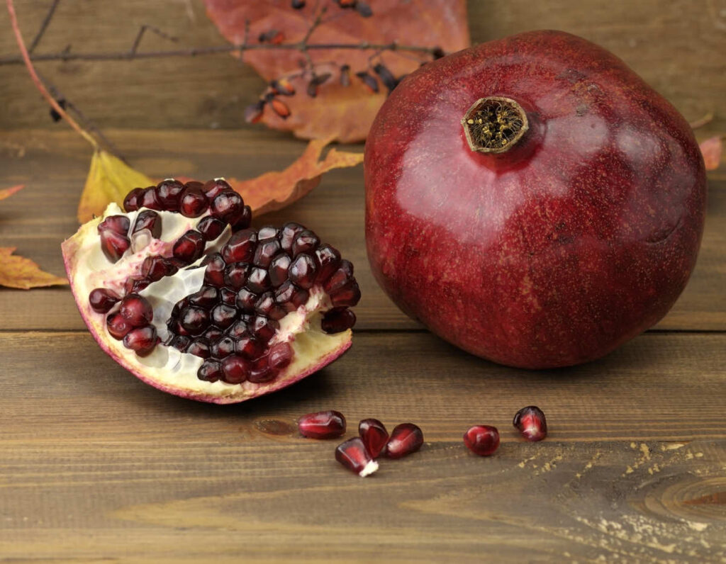 Die Kraft vom Granatapfel 3 Ein ganzer und aufgeschnittener Granatapfel mit sichtbaren Kernen liegt auf einem Holztisch neben Herbstblättern.