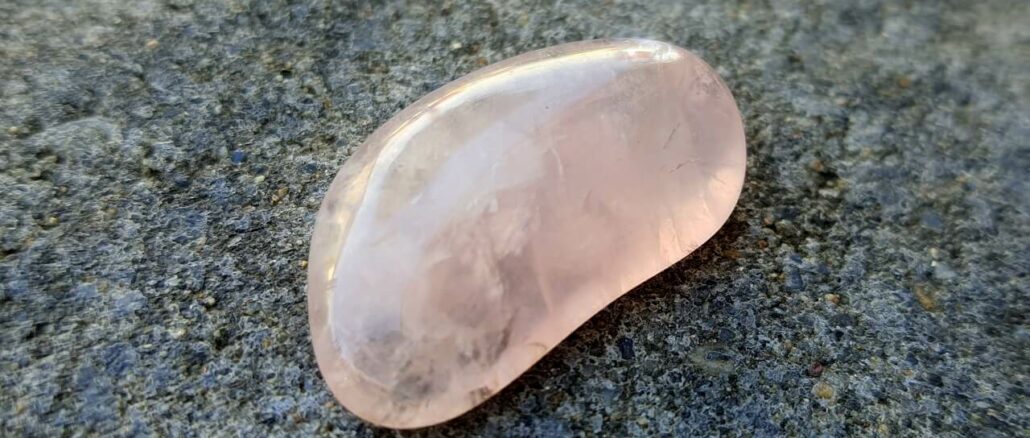 rosenquarz-geschliffen Ein rosa schimmernder Edelstein liegt auf einer rauen grauen Oberfläche.