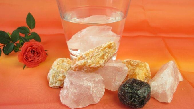 Ein Glas Wasser mit Eiswürfeln, umgeben von Kristallen und einer kleinen roten Rose auf einem orangefarbenen Hintergrund.